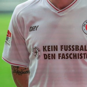 Auf der Brust des weißen Auswärtstrikots des FC St. Pauli ist „Kein Fußball den Faschisten“ zu lesen. Am Arm trägt die Person, deren Kopf nicht zu sehen ist, eine Binde in Regenbogenfarben.