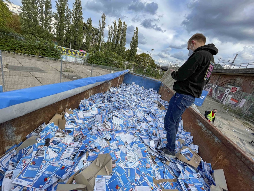 Ein Mann steht in einem Müllcontainer, der mit AfD-Flyern gefüllt ist.