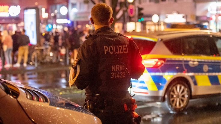Ein Polizist einer Hundertschaft steht nachts neben einem Polizeiauto auf der Zülpicher Straße