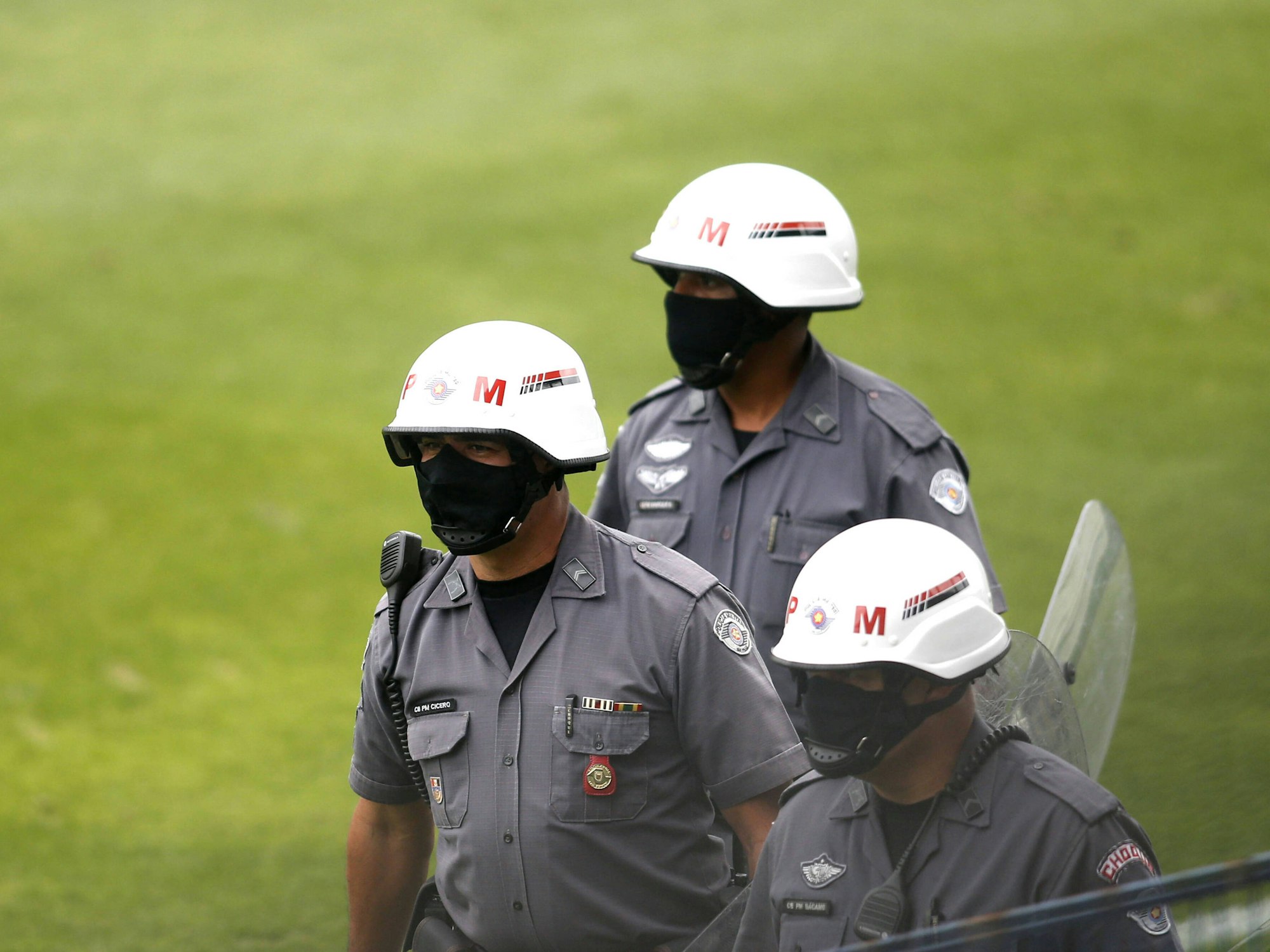 Drei uniformierte Militärpolizisten laufen über das Feld. Sie tragen Helme und Masken. Einer trägt ein Schild in der Hand.