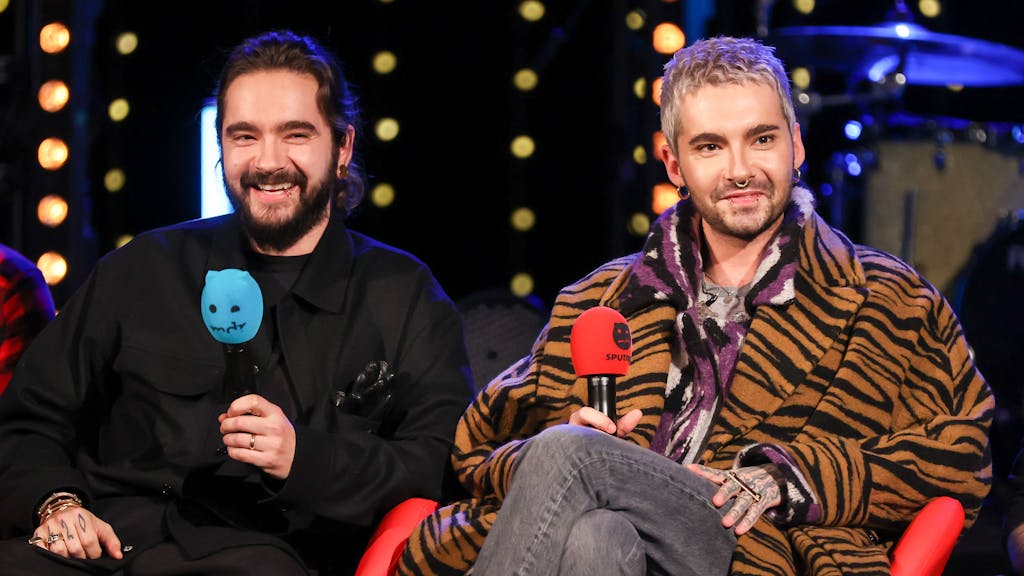 Tom und Bill Kaulitz von der Band Tokio Hotel sitzen während einer Radioshow auf einer Bühne mit jeweils einem Mikrofon in der Hand.