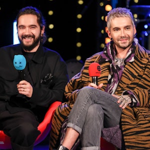 Tom und Bill Kaulitz von der Band Tokio Hotel sitzen während einer Radioshow auf einer Bühne mit jeweils einem Mikrofon in der Hand.