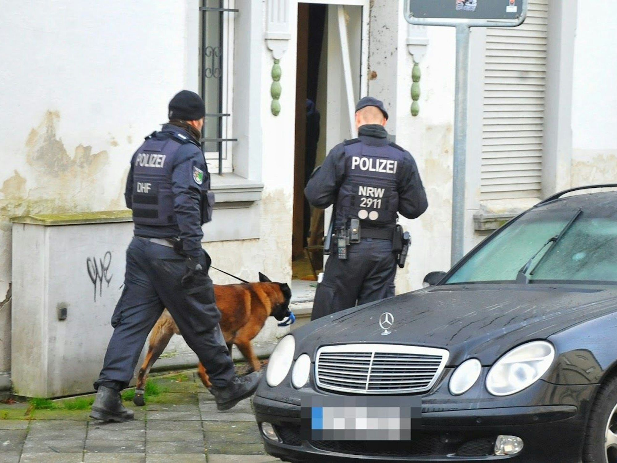 Polizisten und ein Spürhund stehen vor einem Haus.