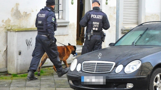 Polizisten und ein Spürhund stehen vor einem Haus.