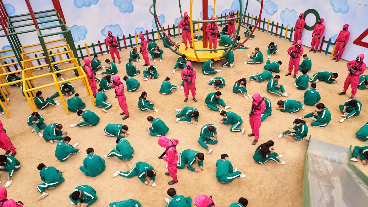 Szene aus der Netflix-Serie „Squid Game“, in der hunderte Erwachsene in Kinderspielen gegeneinander antreten. Die Teilnehmer sitzen auf dem Boden eines nachgebauten Spielplatzes und schnitzen Salzformen aus.
