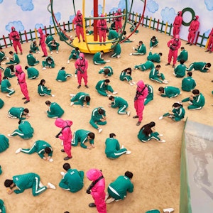 Szene aus der Netflix-Serie „Squid Game“, in der hunderte Erwachsene in Kinderspielen gegeneinander antreten. Die Teilnehmer sitzen auf dem Boden eines nachgebauten Spielplatzes und schnitzen Salzformen aus.
