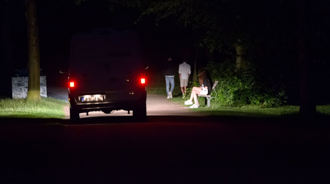 Das Symbolfoto (aufgenommen am 2. Juli 2021) zeigt ein Polizeifahrzeug, welches durch einen dunklen, fast menschenleeren Park fährt, während im Licht der Scheinwerfer vier Jugendliche zu sehen sind.