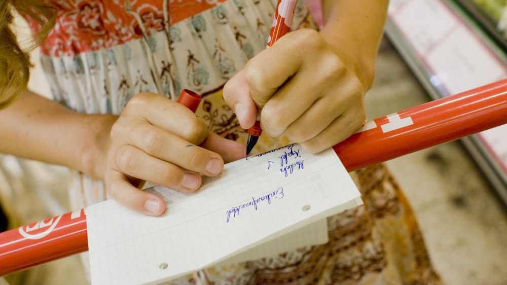 ARCHIV - Ein linkshändiges Mädchen kauft mit einem Einkaufszettel in einem Supermarkt ein und hakt mit einem Stift die Waren ab.