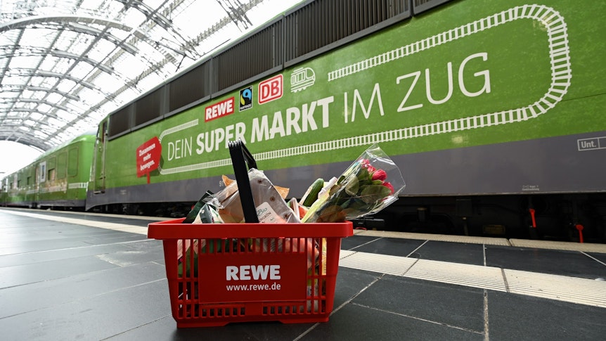 Ein gefüllter Einkaufskorb steht bei der Vorstellung des temporären Projekts "Fairer Supermarkt im Zug" im Frankfurter Hauptbahnhof vor der Lok mit der Aufschrift "Dein Supermarkt im Zug".