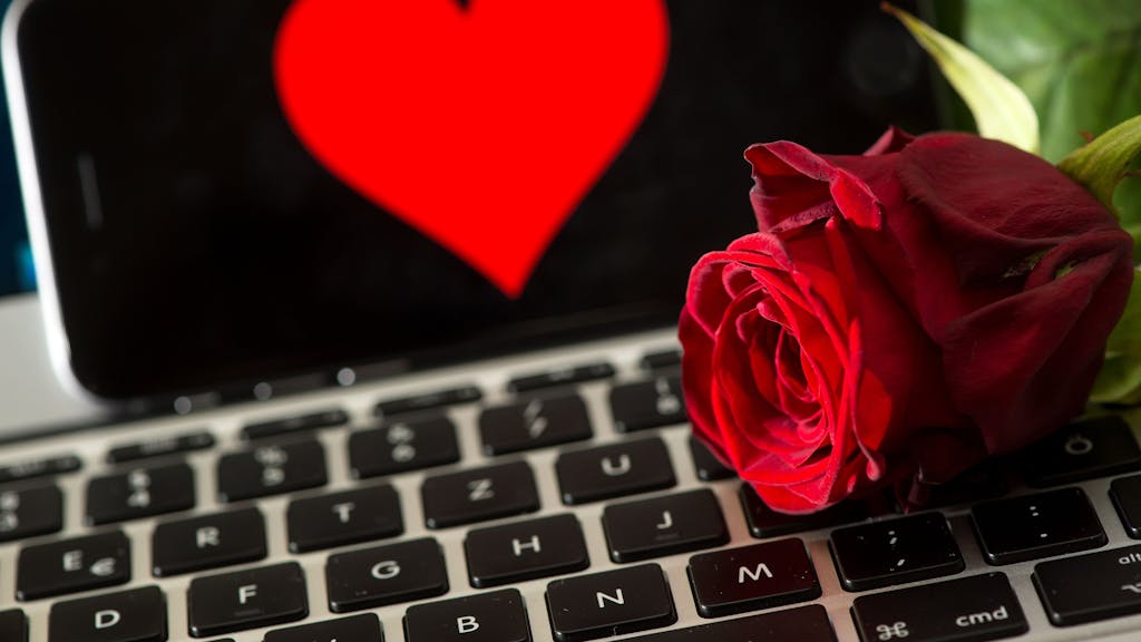 Online-Dating generell ist nicht einfach, doch vor allem gläubige Menschen haben Schwierigkeiten dabei, einen Partner zu finden. Extra Dating-Apps sollen dabei helfen. Das Symbolfoto (aufgenommen am 20. April 2016) zeigt eine Rose, die auf einer Computertastatur liegt. Im Hintergrund ist ein Smartphone zu sehen, auf dem ein Herz abgebildet ist.&nbsp;
