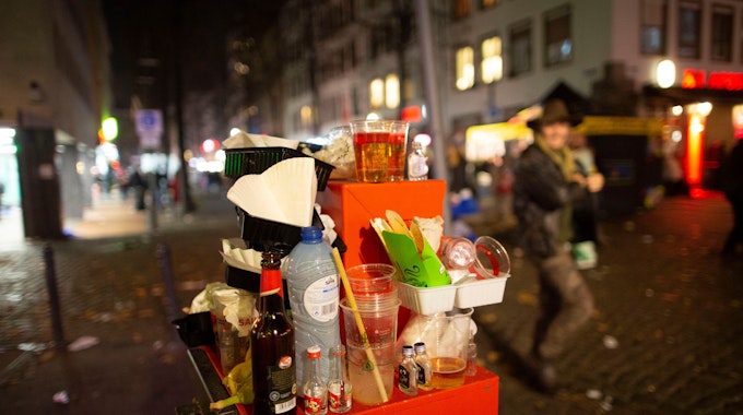 Abfall türmt sich auf einem Mülleimer. Karnevalisten feiern in den Straßen der Kölner Altstadt in den Abend hinein.