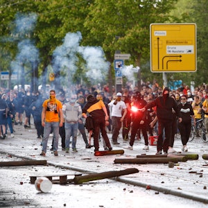 Vermummte Fans von Dynamo Dresden stehen auf der Straße, einer hält ein Bengalo in der Hand.