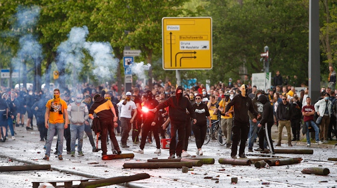 Vermummte Fans von Dynamo Dresden stehen auf der Straße, einer hält ein Bengalo in der Hand.