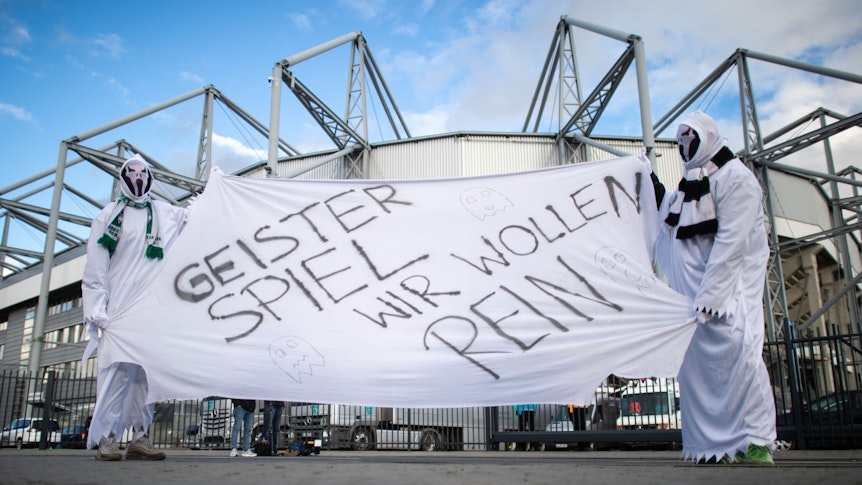 Zwei als Geist verkleidete Gladbach-Fans stehen vor dem Derby gegen den 1. FC Köln am 11. März 2020 vor den Toren des Borussia-Parks. Sie zeigen ein Banner mit der Aufschrift „Geisterspiel, wir wollen rein“.