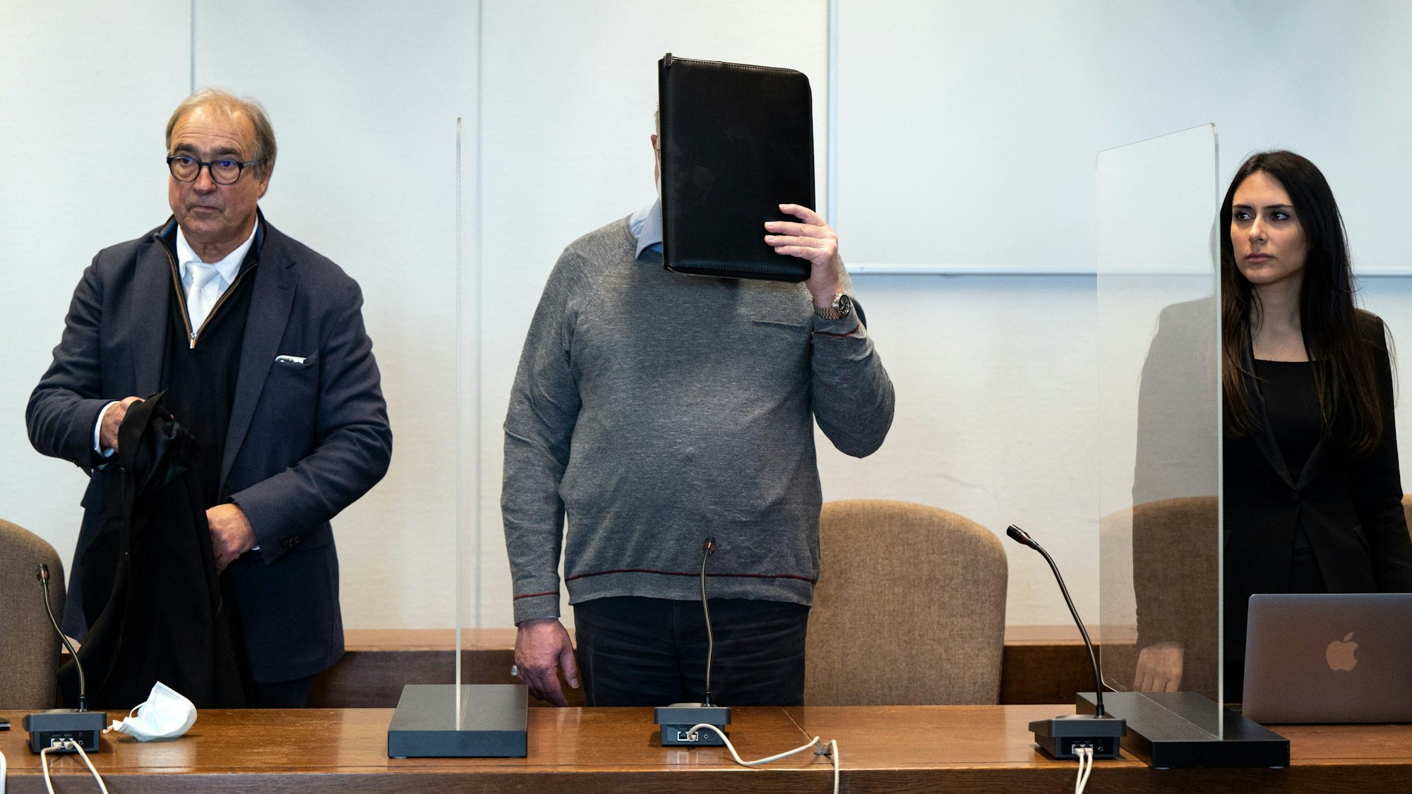 Der angeklagte katholische Priester hält sich im Gerichtssaal eine Mappe vor das Gesicht. Links und rechts stehen sein Anwalt beziehungsweise seine Anwältin.