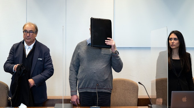 Der angeklagte katholische Priester hält sich im Gerichtssaal eine Mappe vor das Gesicht. Links und rechts stehen sein Anwalt beziehungsweise seine Anwältin.&nbsp;