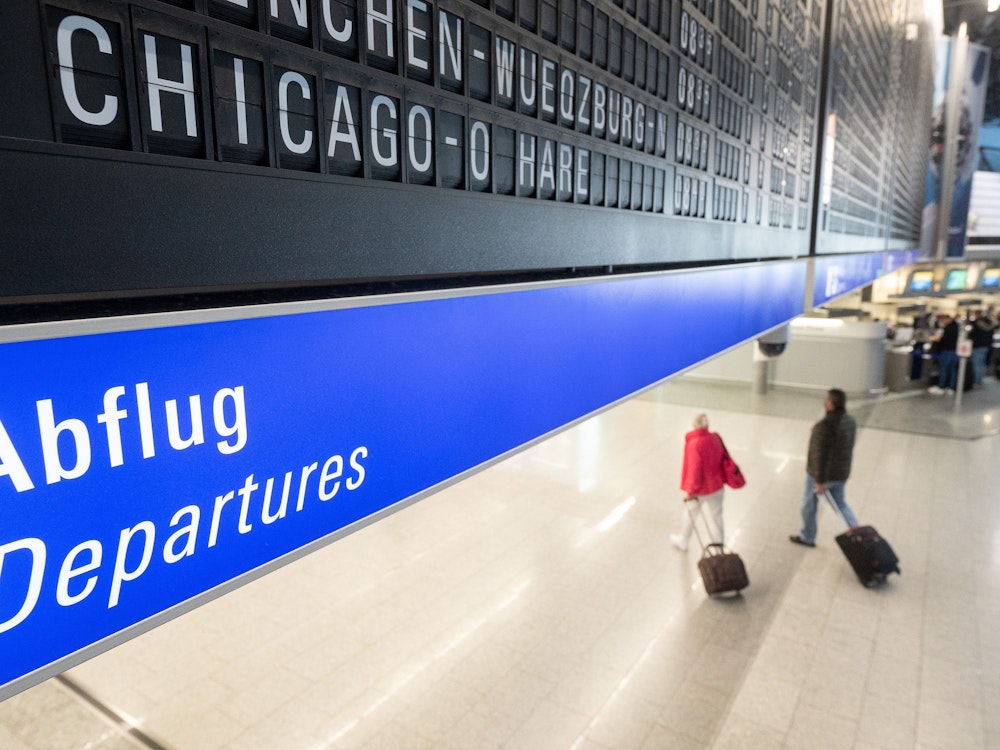 Ein Flug nach Chicago O·Hare in den USA wird auf dem Flughafen Frankfurt auf einer Tafel angezeigt unter der Passagiere mit Koffern entlang gehen.