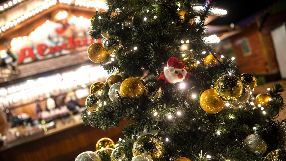Auf dem Symbolfoto sieht man einen geschmückten Weihnachtsbaum, der vor einem beleuchteten Weihnachtsstand leuchtet.