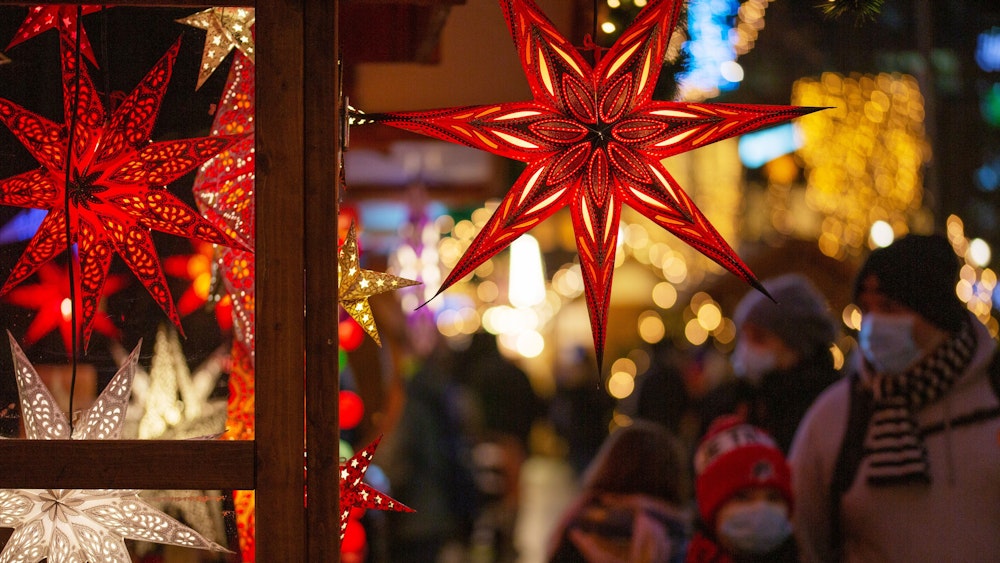 Eine Familie geht mit Mund-Nasen-Schutz auf einem Weihnachtsmarkt an einem Marktstand mit leuchtenden Sternen vorbei.