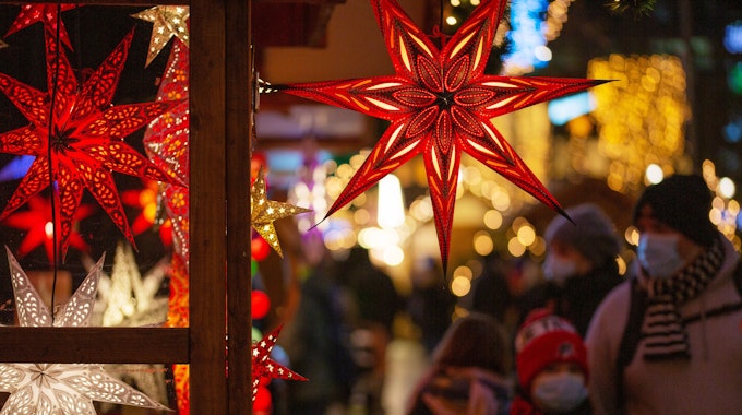Eine Familie geht mit Mund-Nasen-Schutz auf einem Weihnachtsmarkt an einem Marktstand mit leuchtenden Sternen vorbei.