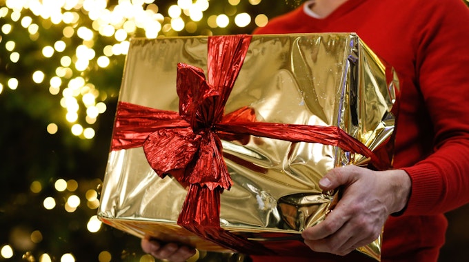 Ein Mann hält ein golden eingepacktes Weihnachtsgeschenk in den Händen. Im Hintergrund ist ein Weihnachtsbaum zu sehen.