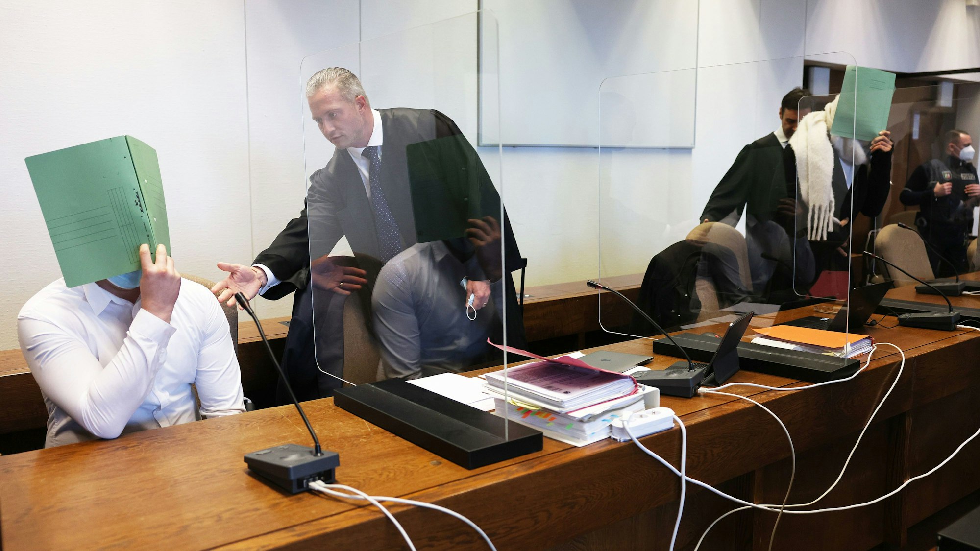 Zwei Angeklagte halten sich neben ihren Verteidigern im Gerichtssaal eine Mappe vor das Gesicht.