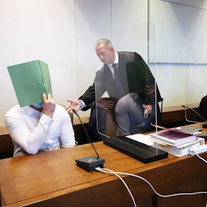 Zwei Angeklagte halten sich neben ihren Verteidigern im Gerichtssaal eine Mappe vor das Gesicht.