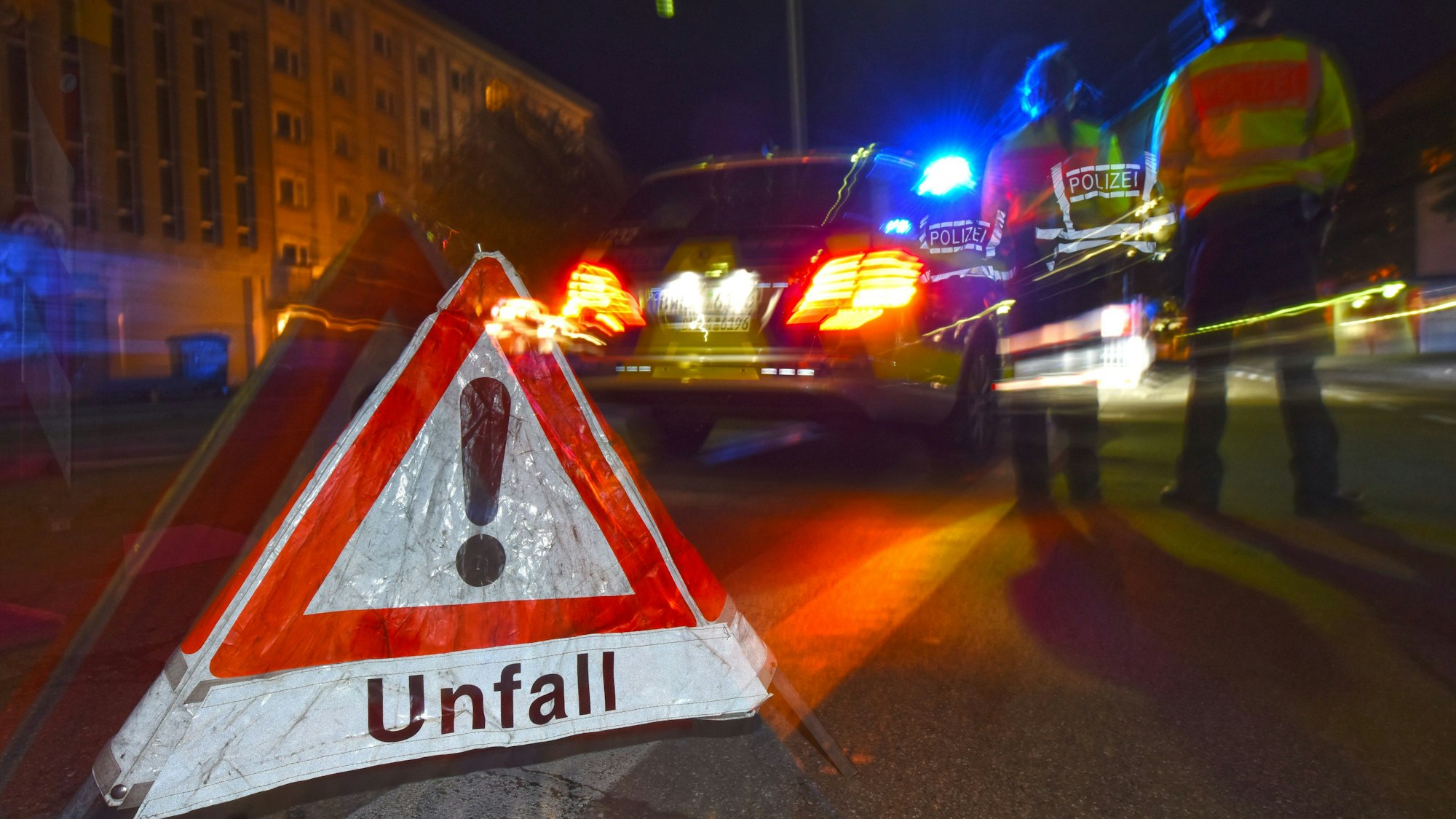 Ein Polizeifahrzeug steht mit Blaulicht auf der Straße. Daneben steht ein Warndreieck mit dem Schriftzug „Unfall“.