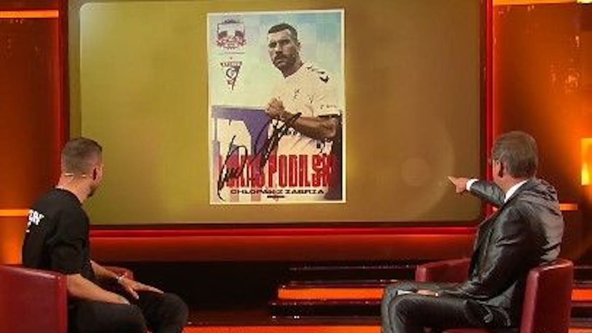 Lukas Podolski schaut mit Günther Jauch auf die Autogrammkarte.