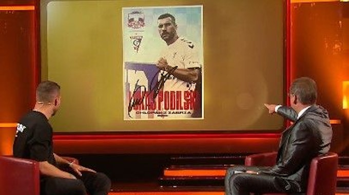 Lukas Podolski schaut mit Günther Jauch auf die Autogrammkarte.