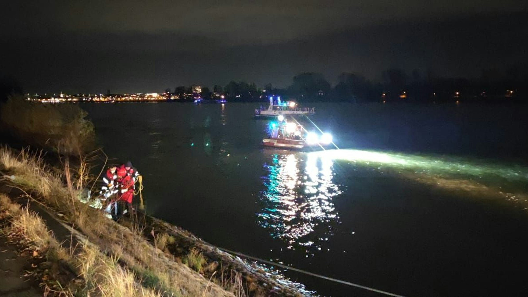 Einsatzkräfte stehen am Rheinufer, auf dem Wasser beleuchtet ein Boot die Wasseroberfläche.