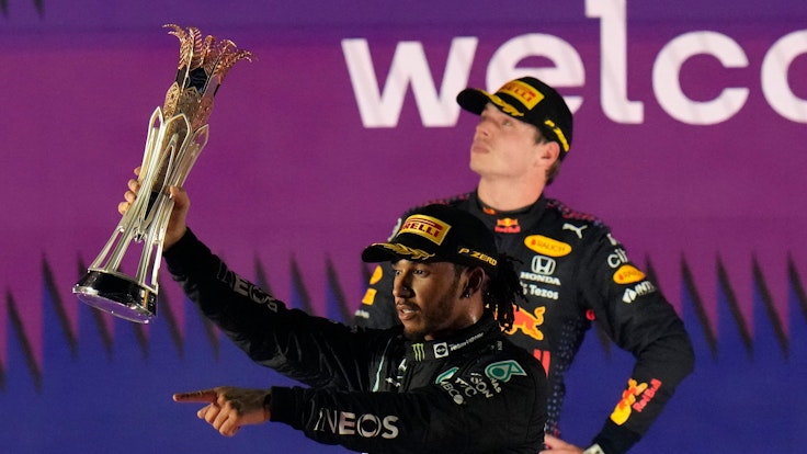 Lewis Hamilton jubelt nach einem Sieg in der Formel 1 mit Pokal, Max Verstappen steht zerknirscht dahinter.