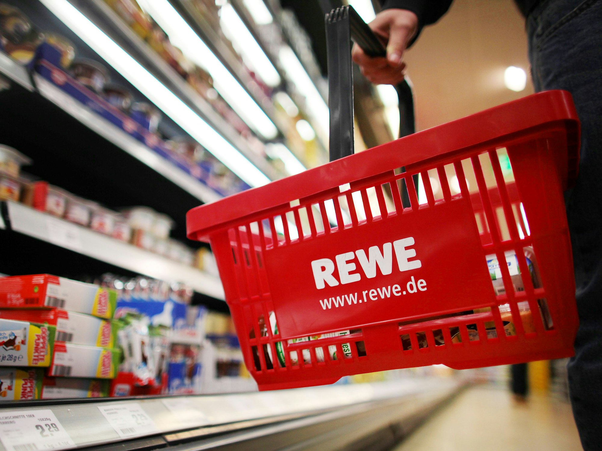Ein Kunde steht mit einem roten Einkaufskorb mit der Aufschrift „Rewe“ vor einem Einkaufsregal.