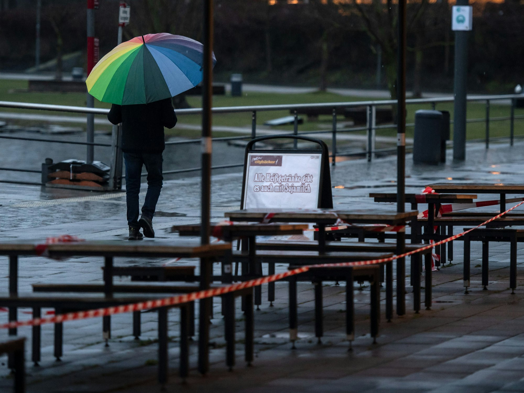 Ein Passant geht unter einem bunten Regenschirm an der Uferprommenade des Phoenix Sees an den wegen der Corona-Pandemie gesperrten Tischen vor einer Bäckerei vorbei.