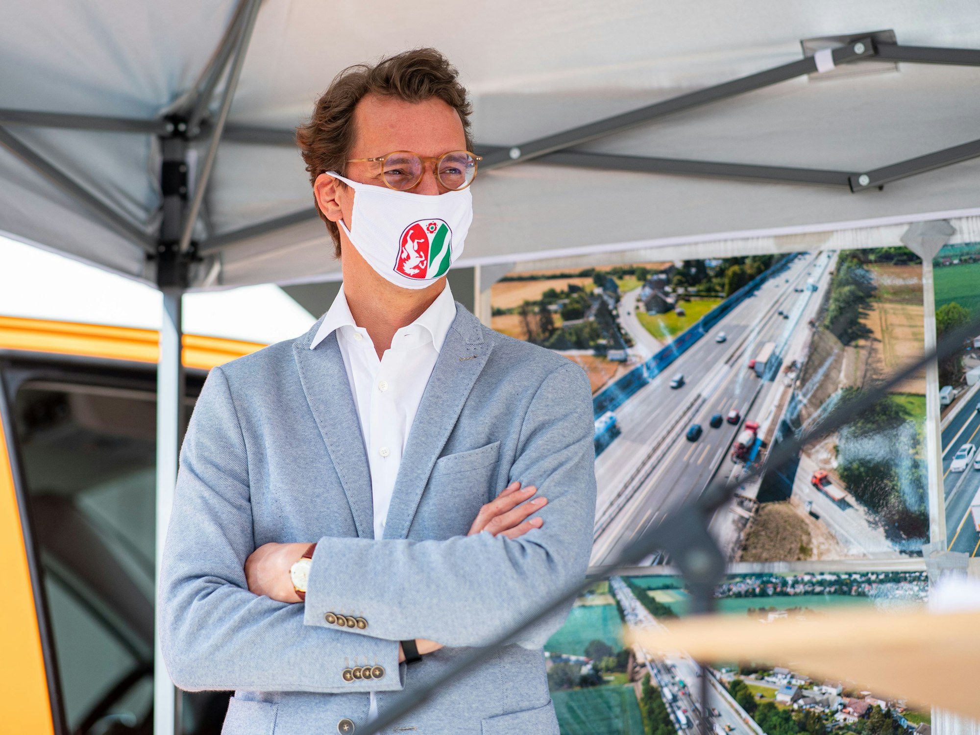 Hendrik Wüst steht mit einer Mund- und Nasenmaske vor einem Bildschirm.