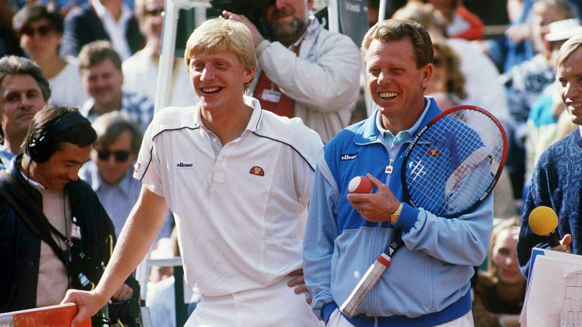Boris Becker und Günther Bosch stehen auf dem Tennisplatz. Becker lehnt sich auf das Netz, Bosch hält einen Schläger und einen Ball.