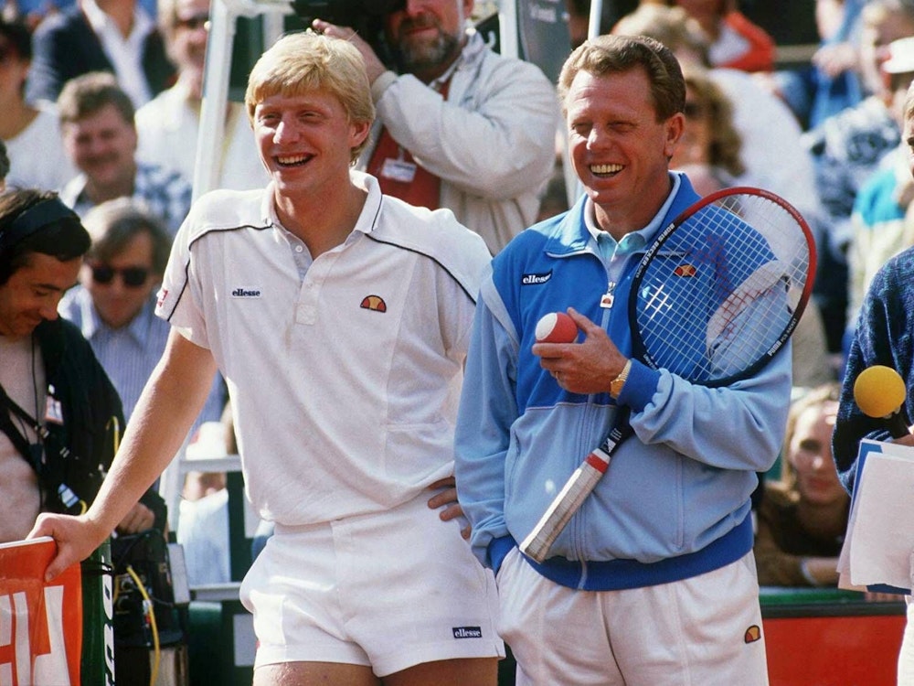 Boris Becker und Günther Bosch stehen auf dem Tennisplatz. Becker lehnt sich auf das Netz, Bosch hält einen Schläger und einen Ball.