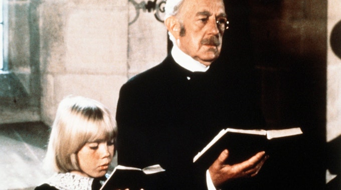 Der alte Earl of Dorincourt (Alec Guinness) steht neben seinem Enkel Ceddie (Ricky Schroder). Beide halten ein Gesangsbuch in der Hand.&nbsp;