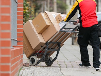 Ein Paketzusteller geht mit einer Sackkarre voller Pakete zu einem Haus.
