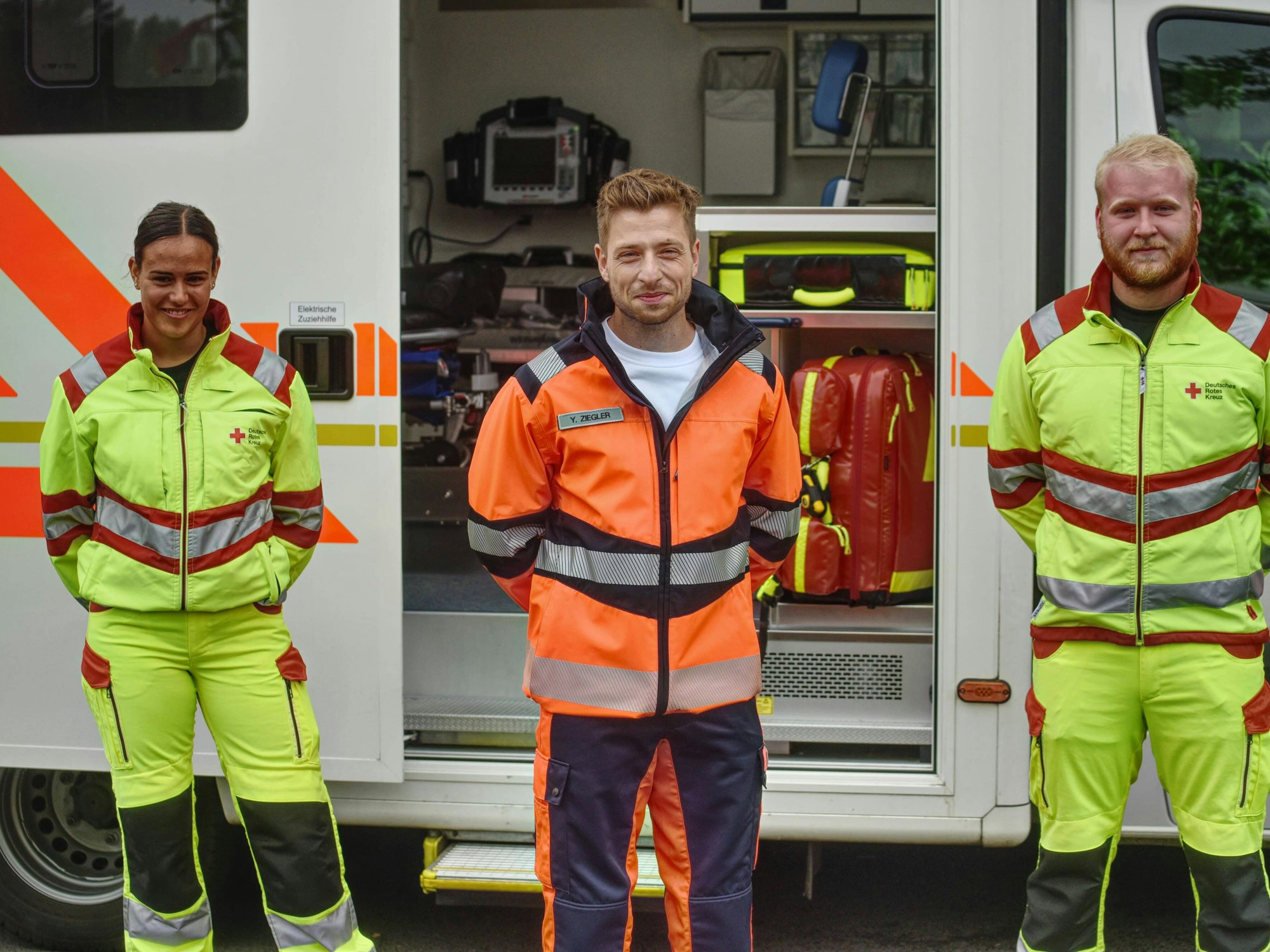 Dominik Flade steht in Arbeitskleidung des Notfallsanitäters zwischen zwei Auszubildenden vor einem RTW.