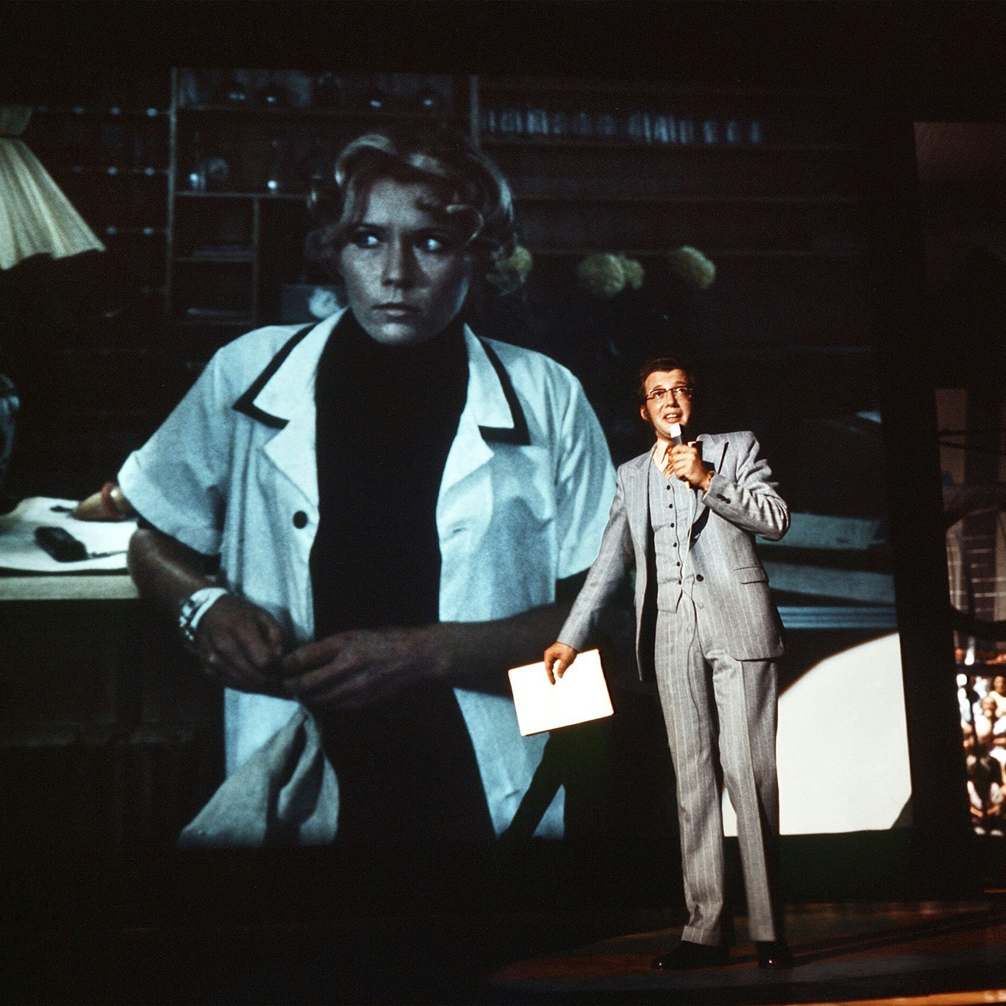 TV-Film „Das Millionenspiel“ (1970): Dieter Thomas Heck als Moderator Tilo Uhlenhorst im Studio spricht mit einer Hotelangestellten (auf der Videowand im Hintergrund)