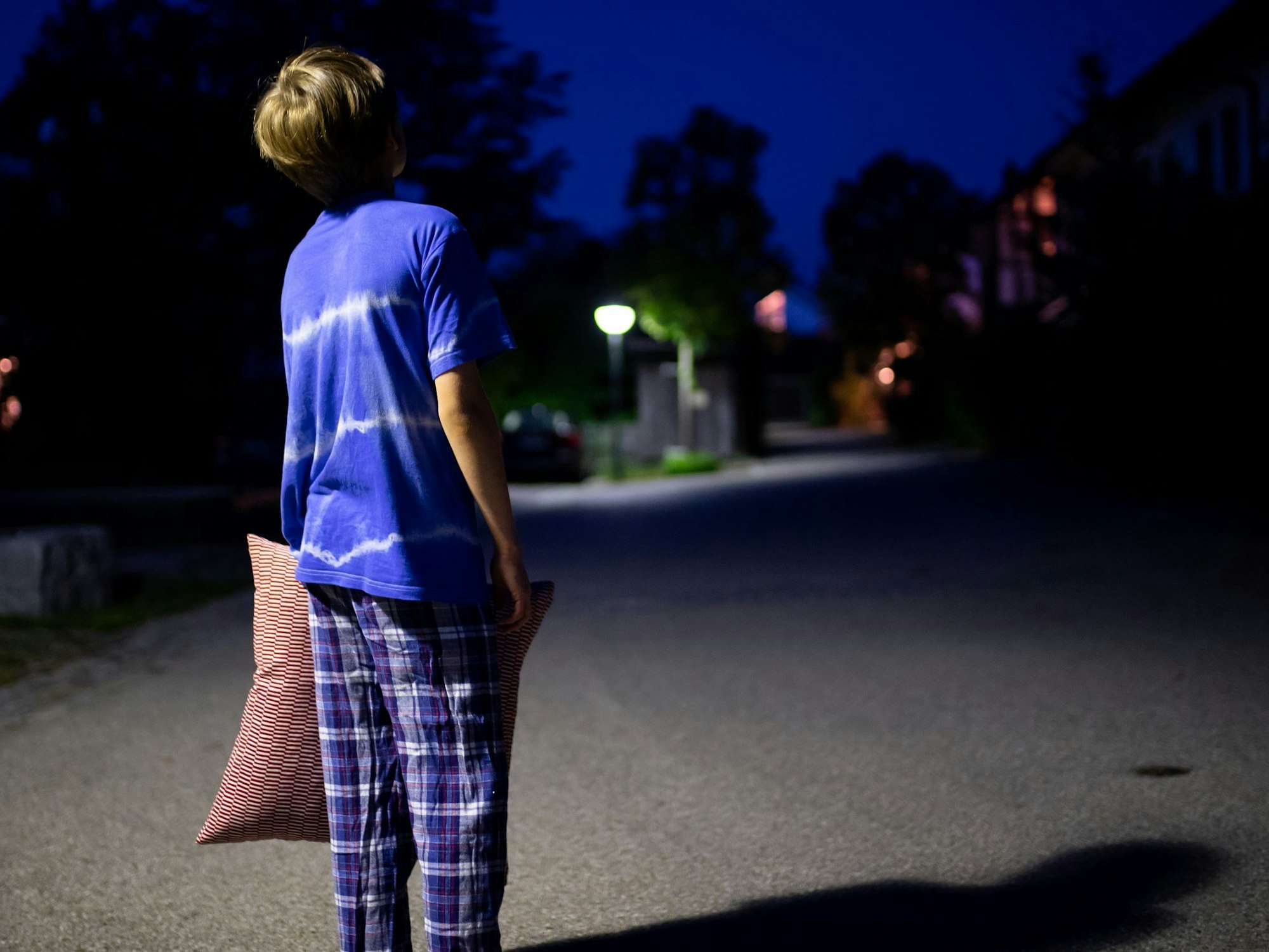 Ein Junge im Schlafanzug steht nachts mit einem Kissen in der Hand auf einer Straße.