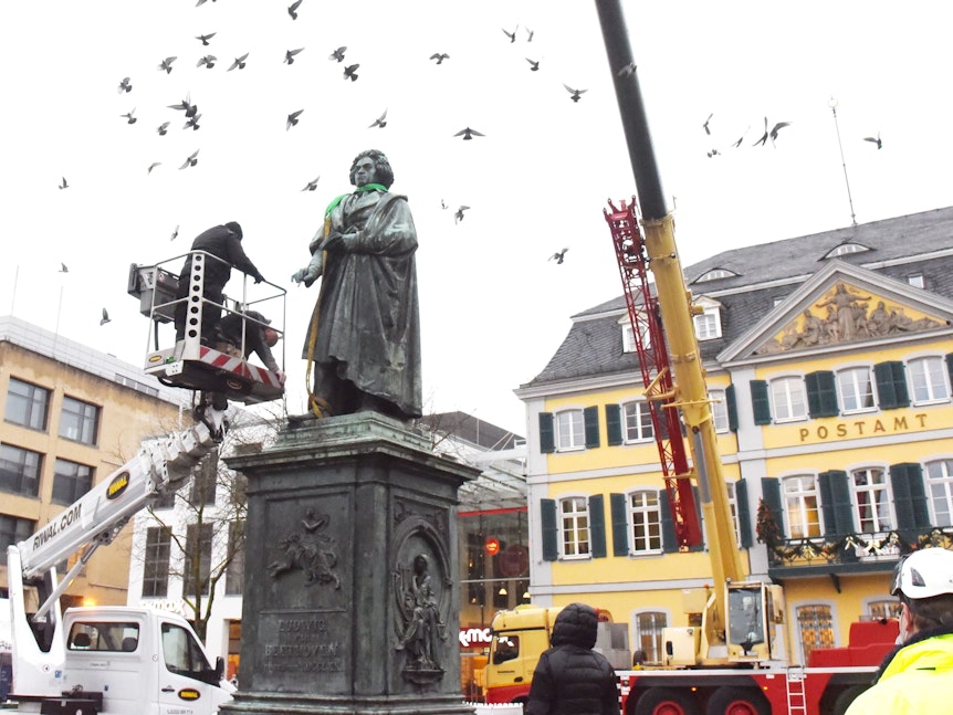 Tauben fliegen um das Beethoven-Denkmal, davor steht ein Mann auf einer Arbeitsbühne.