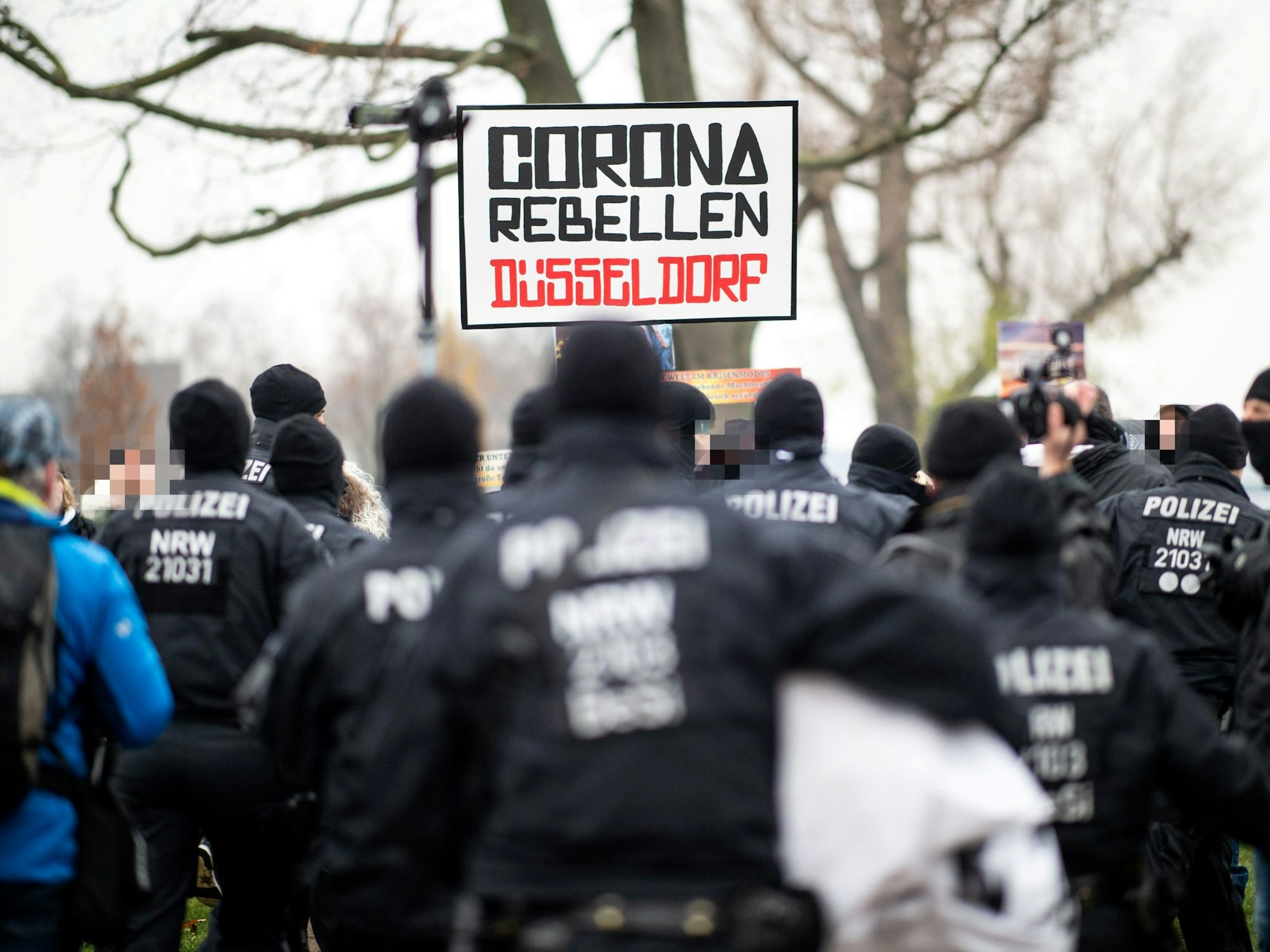 Demonstranten halten bei einer Demonstration am Rheinufer ein Schild mit der Aufschrift „Corona Rebellen Düsseldorf“ in die Höhe, davor stehen Polzisten. D