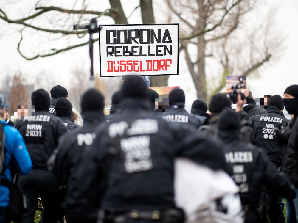 Demonstranten halten bei einer Demonstration am Rheinufer ein Schild mit der Aufschrift „Corona Rebellen Düsseldorf“ in die Höhe, davor stehen Polzisten.