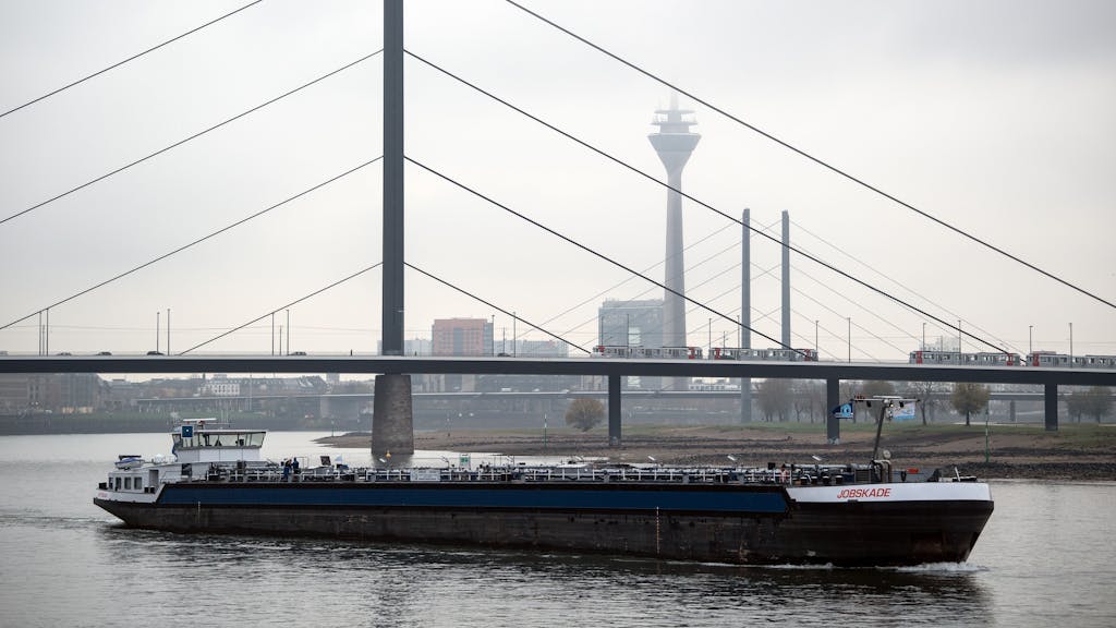 Ein Frachtschiff fährt auf dem Rhein vor der Kulisse der Rheinbrücken und des Rheinturms, der im Dunst beinahe verschwindet.