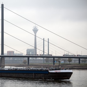 Ein Frachtschiff fährt auf dem Rhein vor der Kulisse der Rheinbrücken und des Rheinturms, der im Dunst beinahe verschwindet.