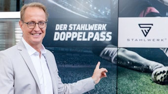 Florian König steht für ein Pressefoto vor einem Bildschirm mit dem Doppelpass-Schriftzug. 