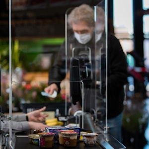 Ein Kunde mit Mundschutz bezahlt an der Kasse seinen Einkauf. An einer Kasse in einem Supermarkt wird die Kassiererin mit einer Plexiglasscheibe, einem sogenannten "Spuckschutz", geschützt.