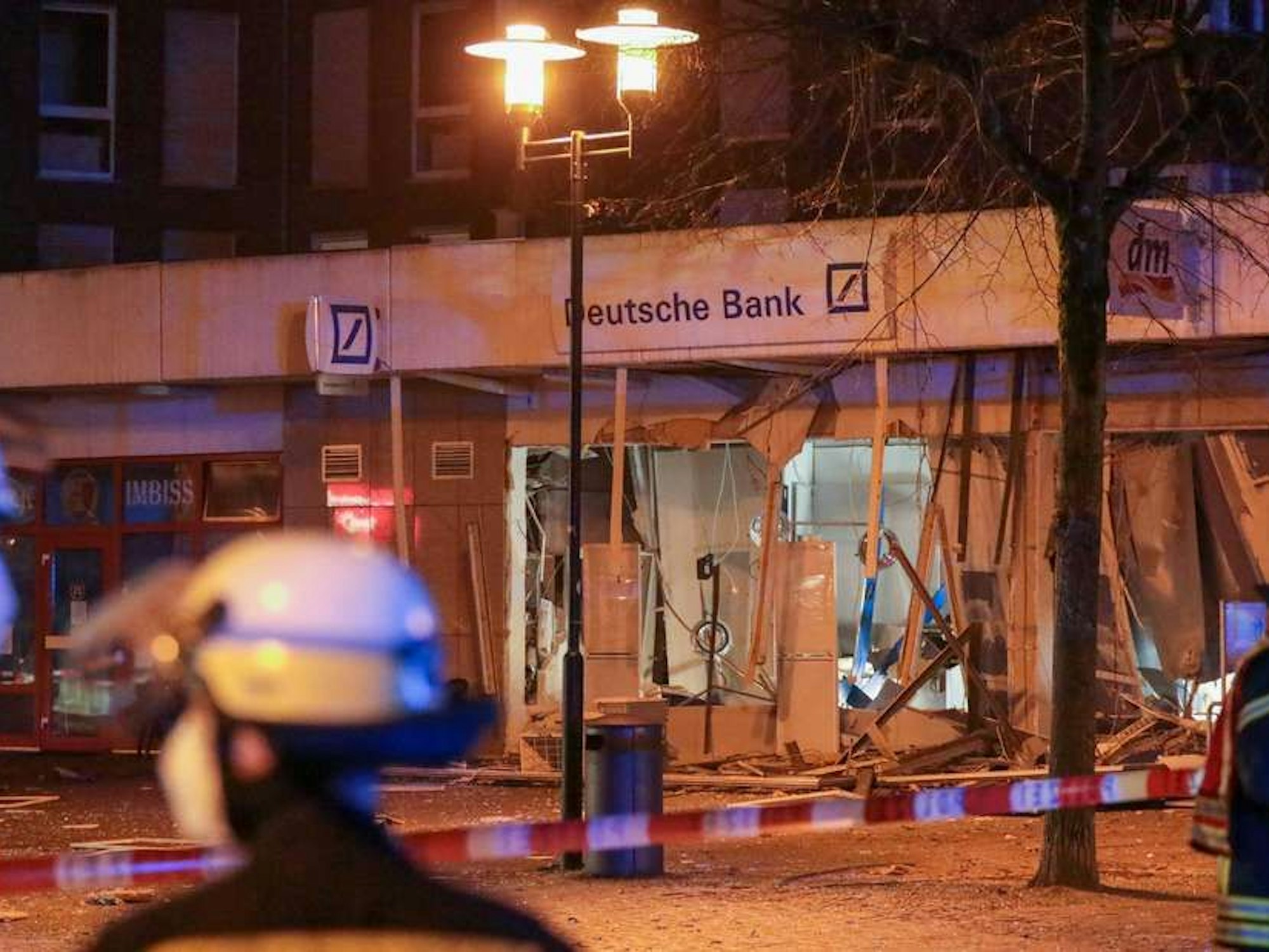 Einsatzkräfte der Feuerwehr stehen vor einer durch eine Explosion stark beschädigten Deutsche Bank-Filiale.